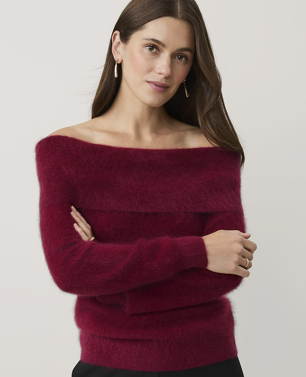 A woman wearing a burgundy off-the-shoulder sweater stands with her arms crossed against a plain, light-colored background.