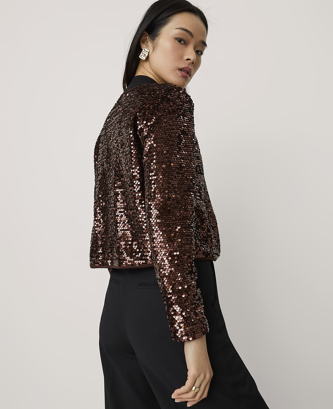A woman wearing a shimmering brown sequined jacket and black trousers stands against a plain, light-colored background.