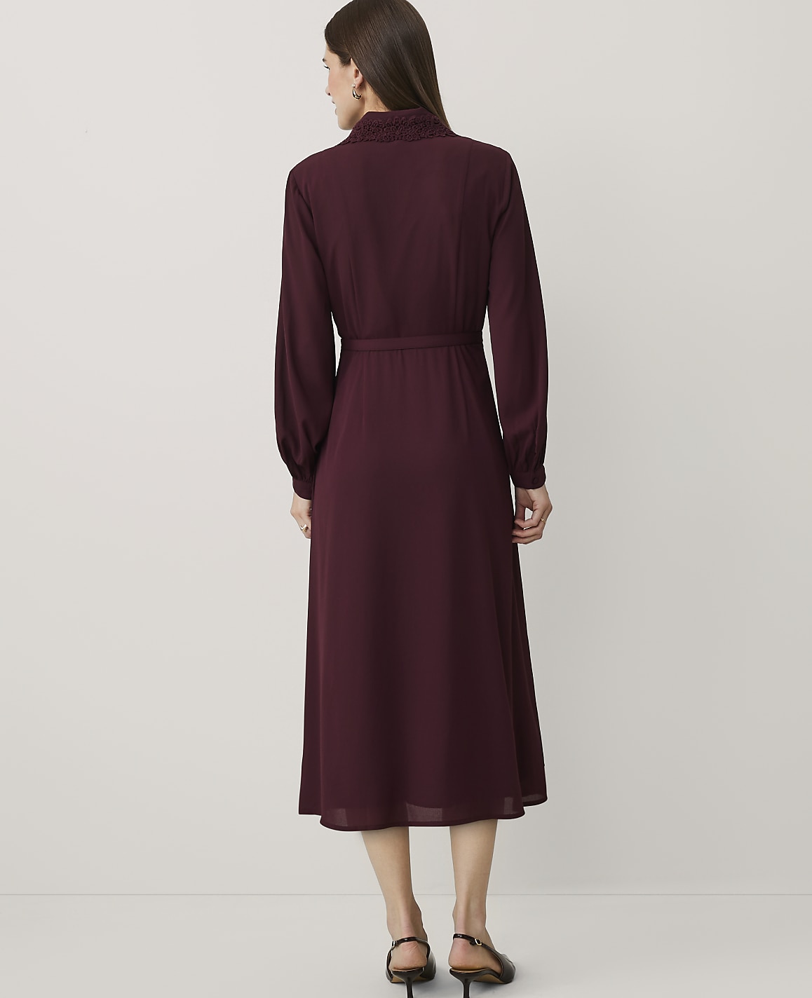 A woman stands with her back to the camera wearing a long-sleeved burgundy dress against a plain white background.
