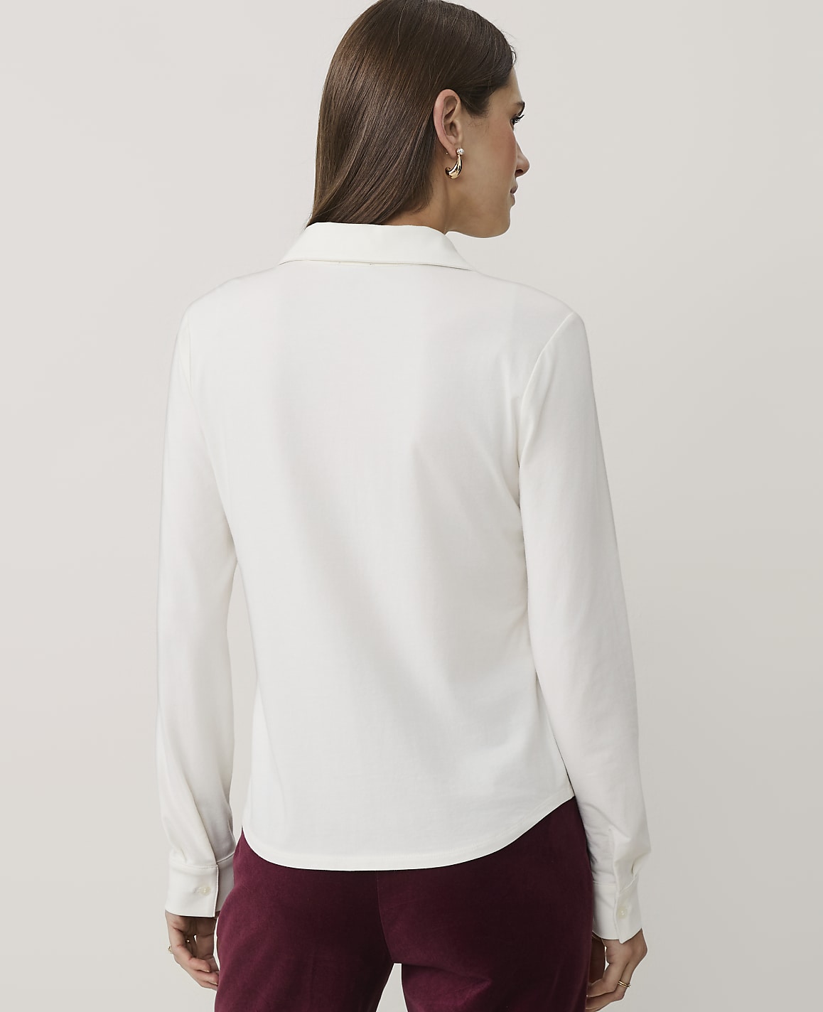 A woman wearing a white long-sleeved shirt and burgundy trousers stands with her back to the camera against a plain light gray background.