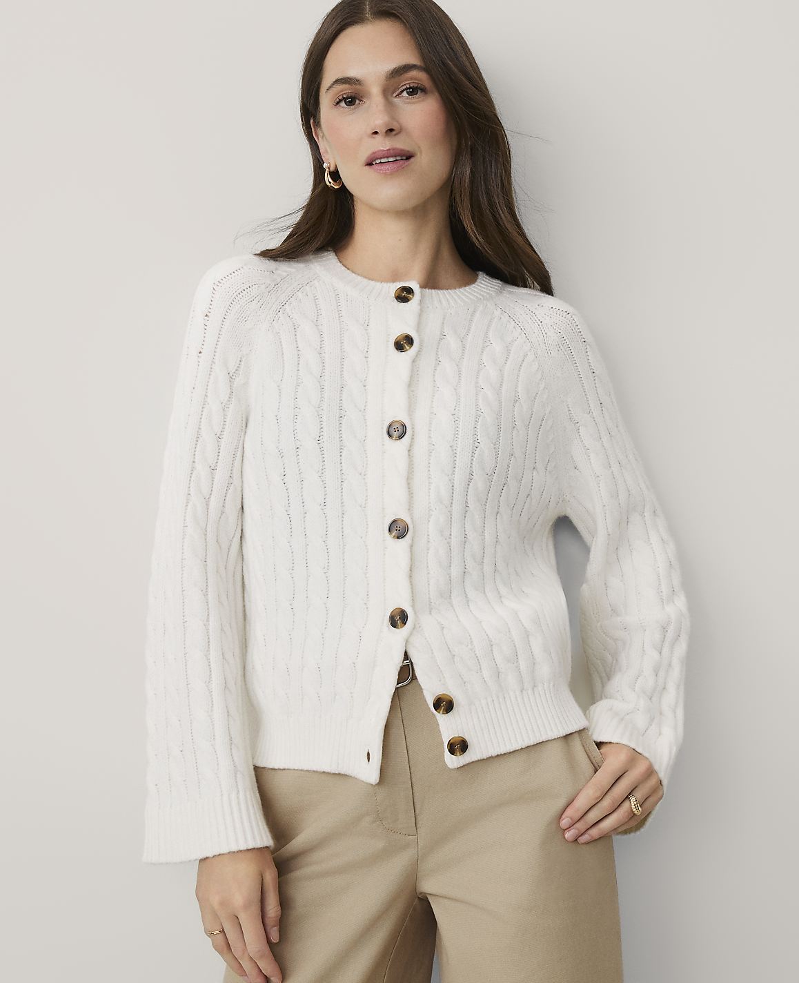 A woman wearing a white cable-knit cardigan and khaki pants stands against a plain, light-colored wall.