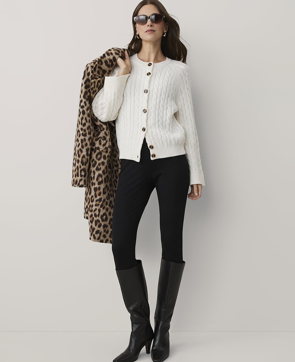 A woman wearing a white cable-knit cardigan, black pants, and black boots stands against a plain white background while holding a leopard-print coat over her shoulder.