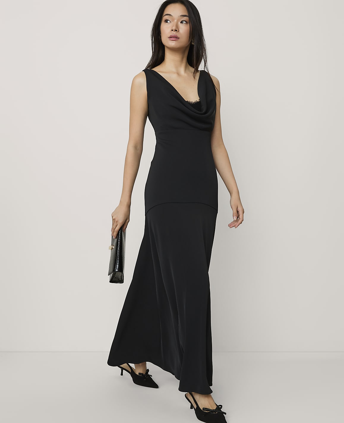 A woman wearing a long black cowl-neck dress and black heels stands against a plain, light-colored background.