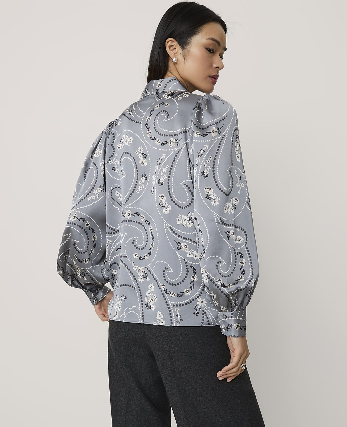 A woman wearing a grey paisley print blouse and dark trousers stands against a plain light grey background.