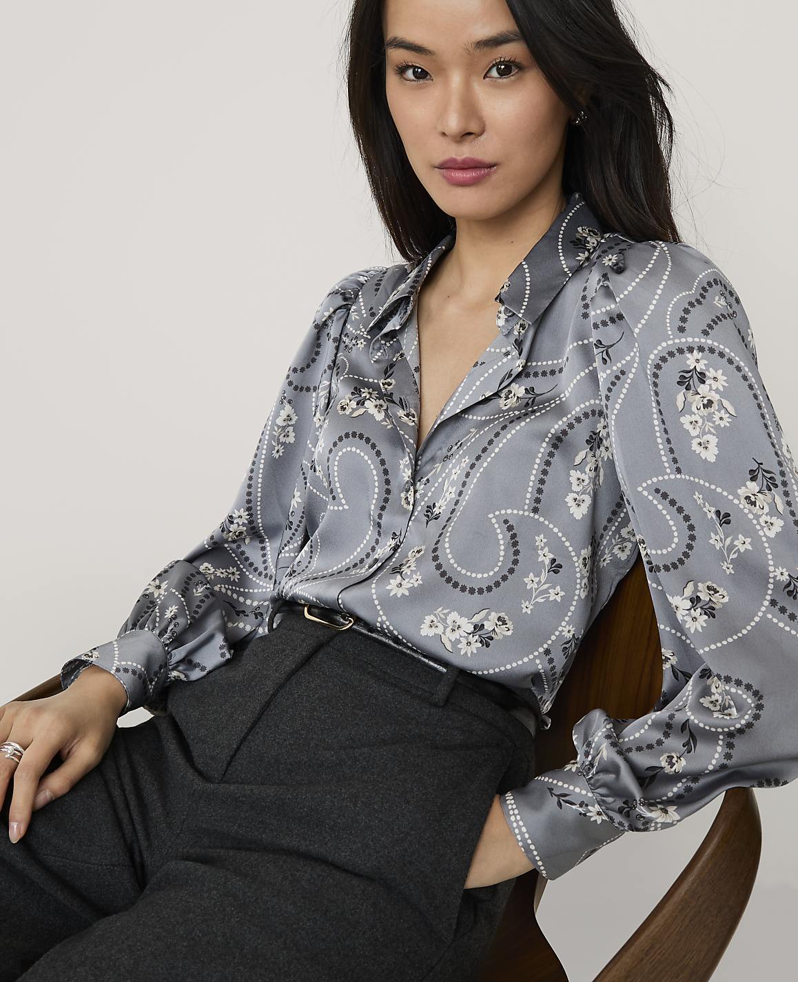 A woman wearing a grey paisley print blouse and dark trousers sits in a wooden chair against a plain light background.