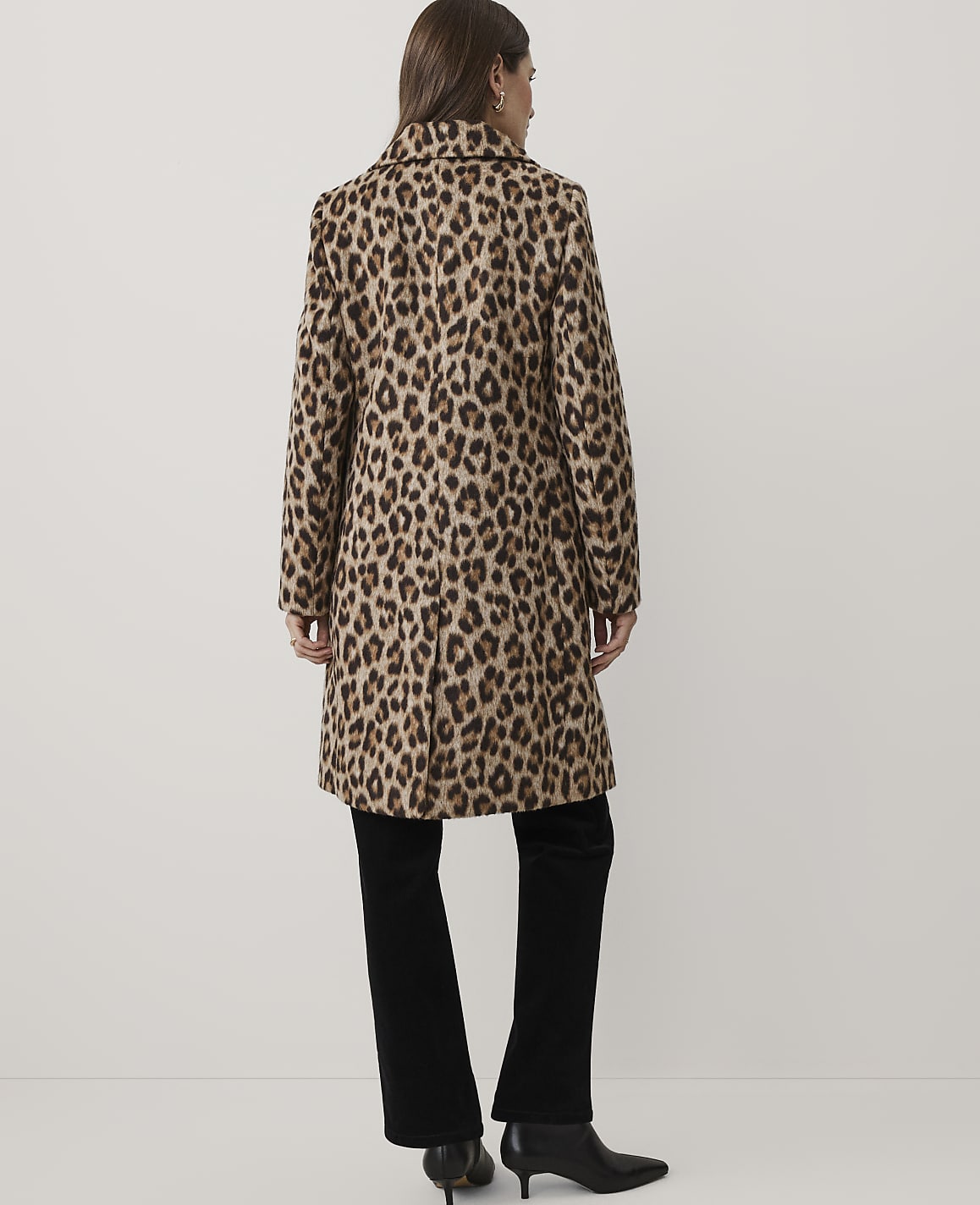 A woman wearing a leopard-print coat and black trousers stands with her back to the camera against a plain white background.