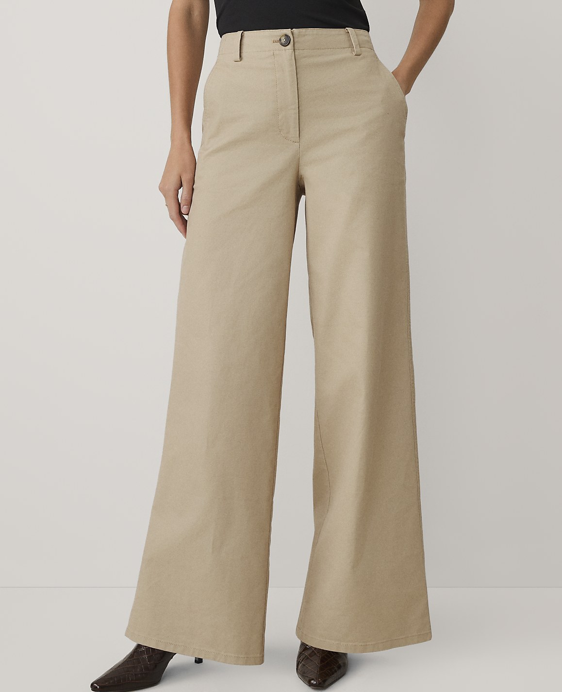A person wears wide-leg beige trousers and dark shoes against a plain light gray background.