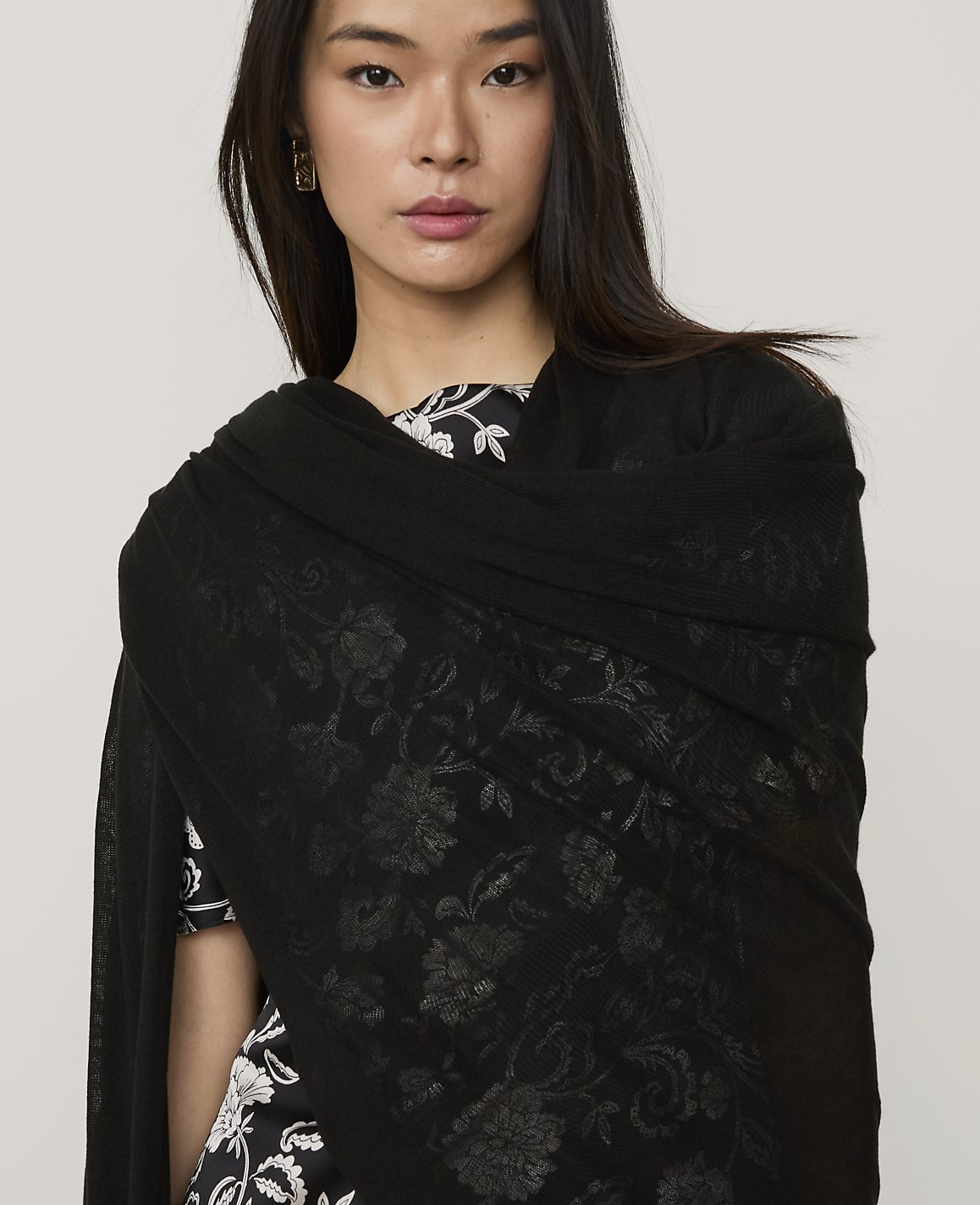 A woman wearing a black floral shawl and a patterned dress stands against a plain, light gray background.