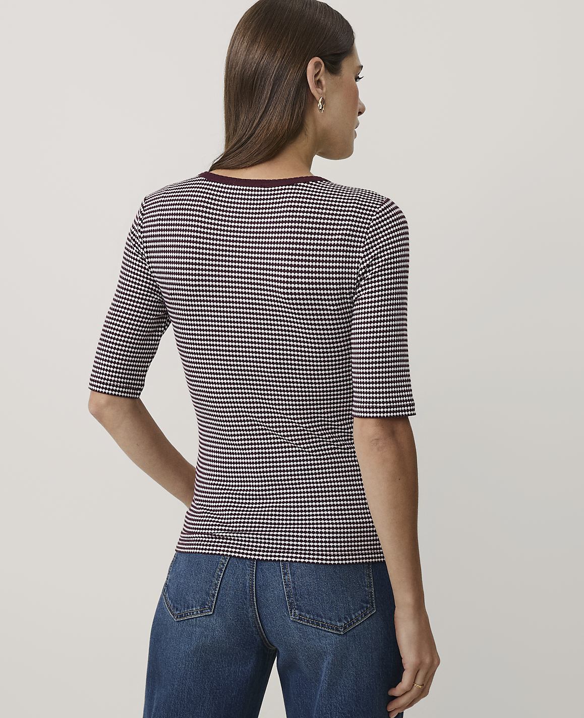 A woman wearing a maroon and white checkered top and blue jeans stands against a plain light gray background.