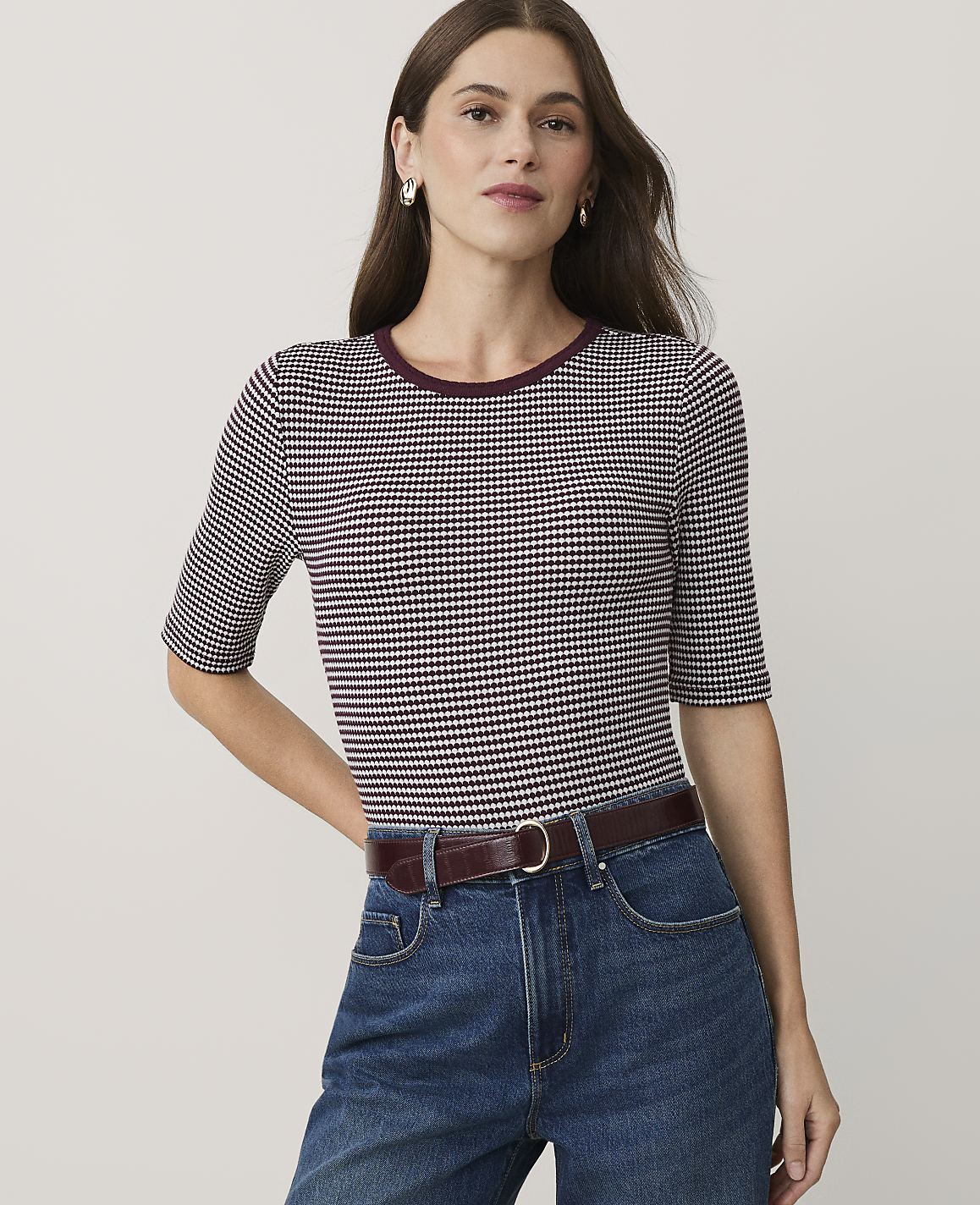 A woman wearing a purple and white patterned top and blue jeans stands against a plain, light-colored background.
