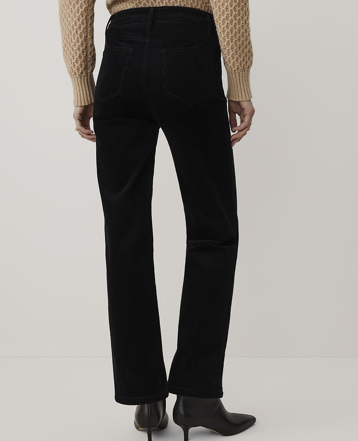 A person wearing a tan knit sweater, black corduroy pants, and black pointed-toe heels stands against a plain white background.