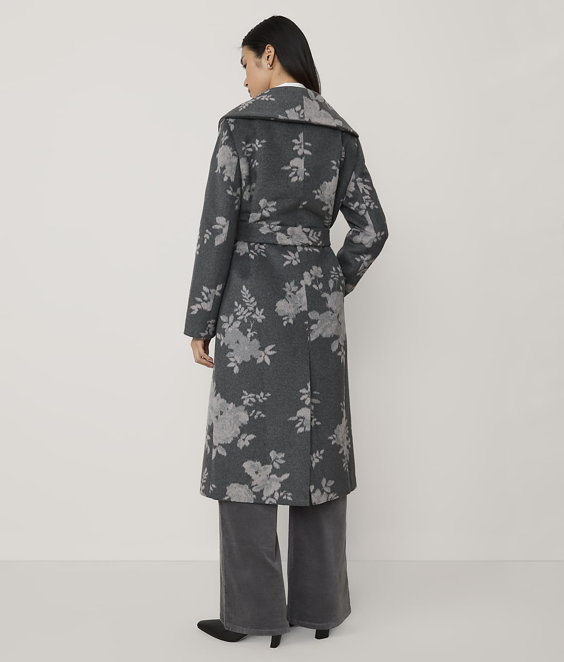 A woman stands with her back to the camera wearing a long, grey floral-patterned coat and wide-leg trousers against a plain, light-colored background.