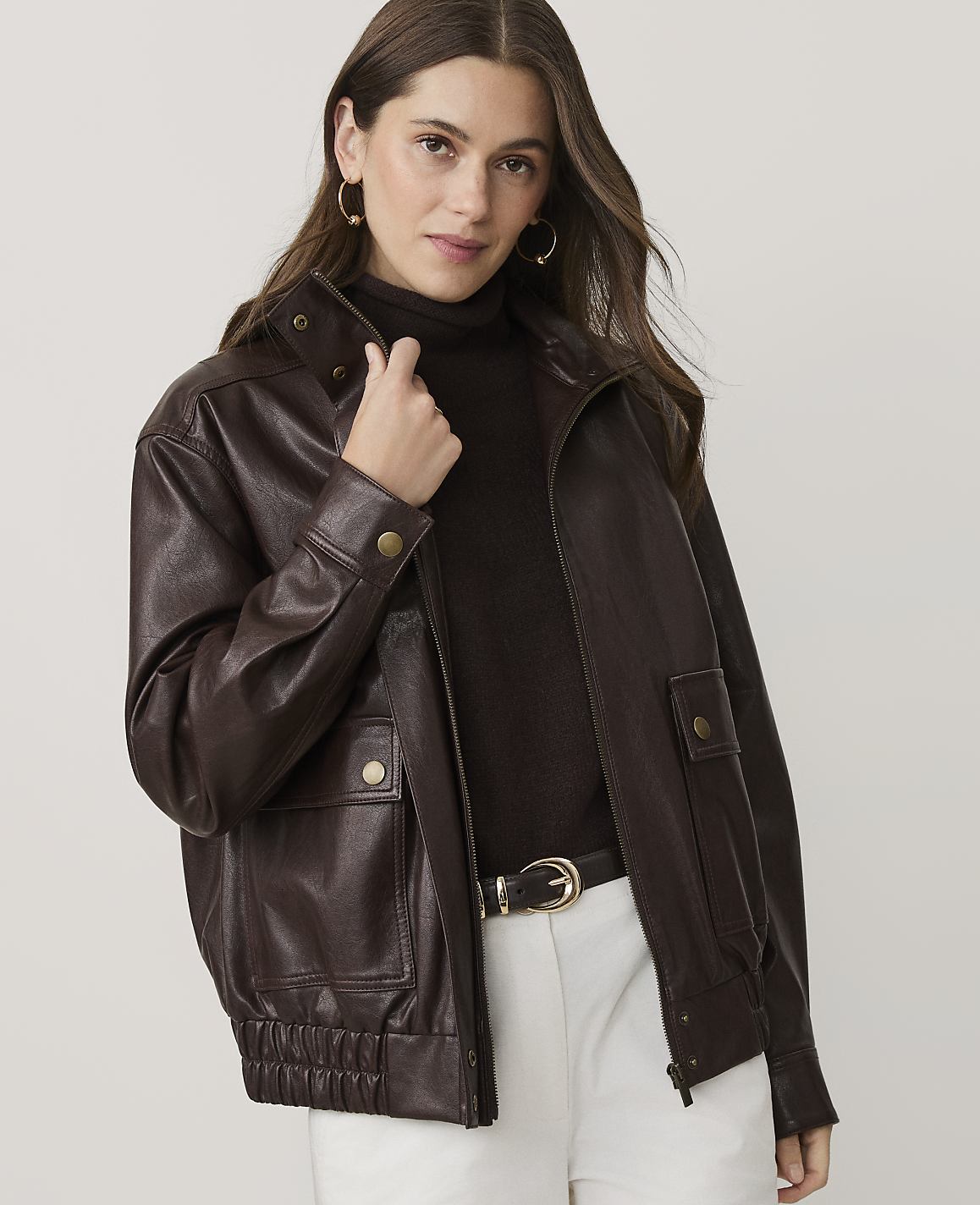 A woman wearing a dark brown leather jacket, a brown turtleneck, and white pants stands against a plain, light-colored background.
