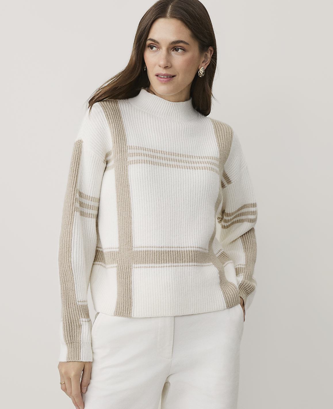 A woman wearing a white and beige plaid sweater and white pants stands against a plain, light-colored background.
