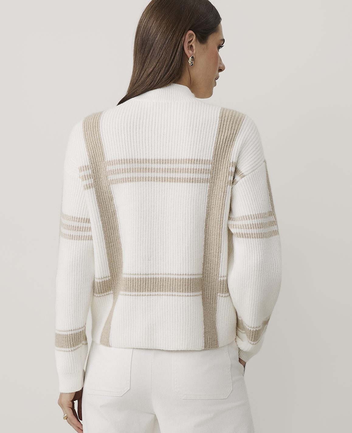 A woman wearing a white and beige plaid sweater and white pants stands against a plain, light-colored background.