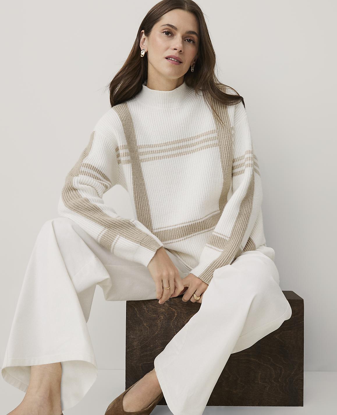 A woman wearing a white and beige patterned sweater and white trousers sits on a dark wooden block against a plain light grey background.