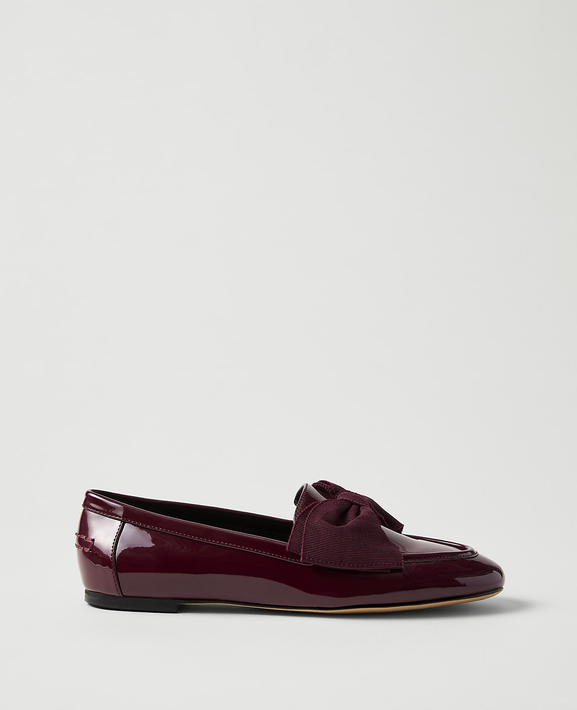 A burgundy patent leather flat shoe with a bow sits in the foreground against a plain, light gray background.