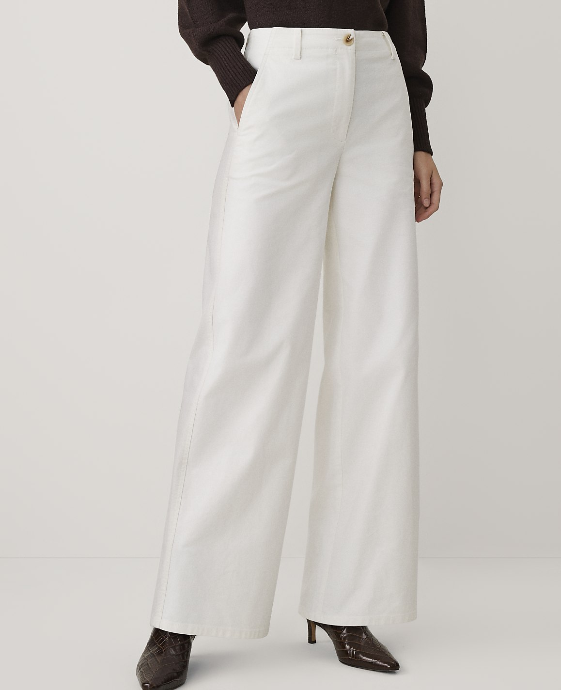 A person wearing a brown sweater, wide-leg white trousers, and dark brown heels stands against a plain, light-colored background.