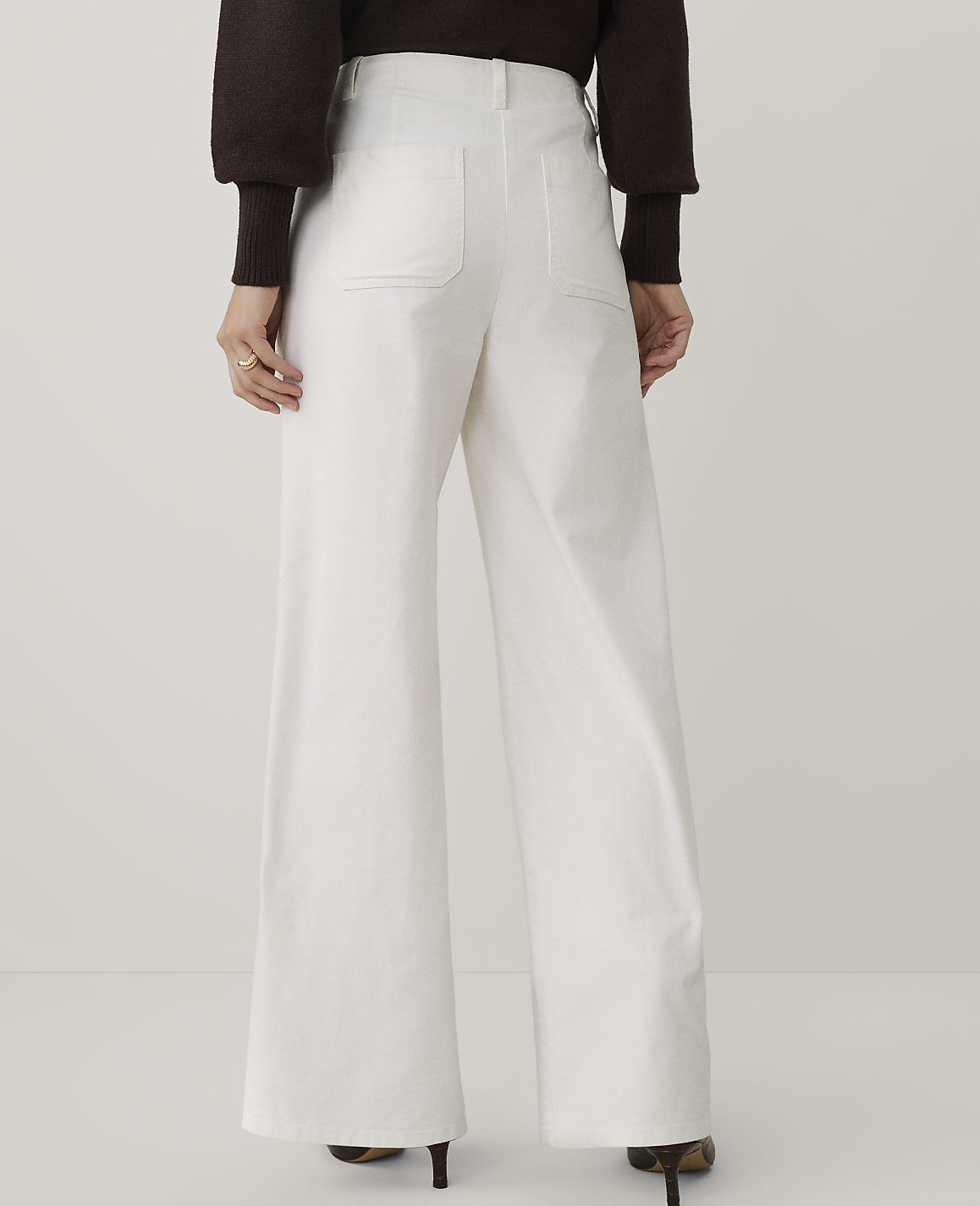 A person wearing a dark brown sweater and wide-leg white trousers stands against a plain light gray background.