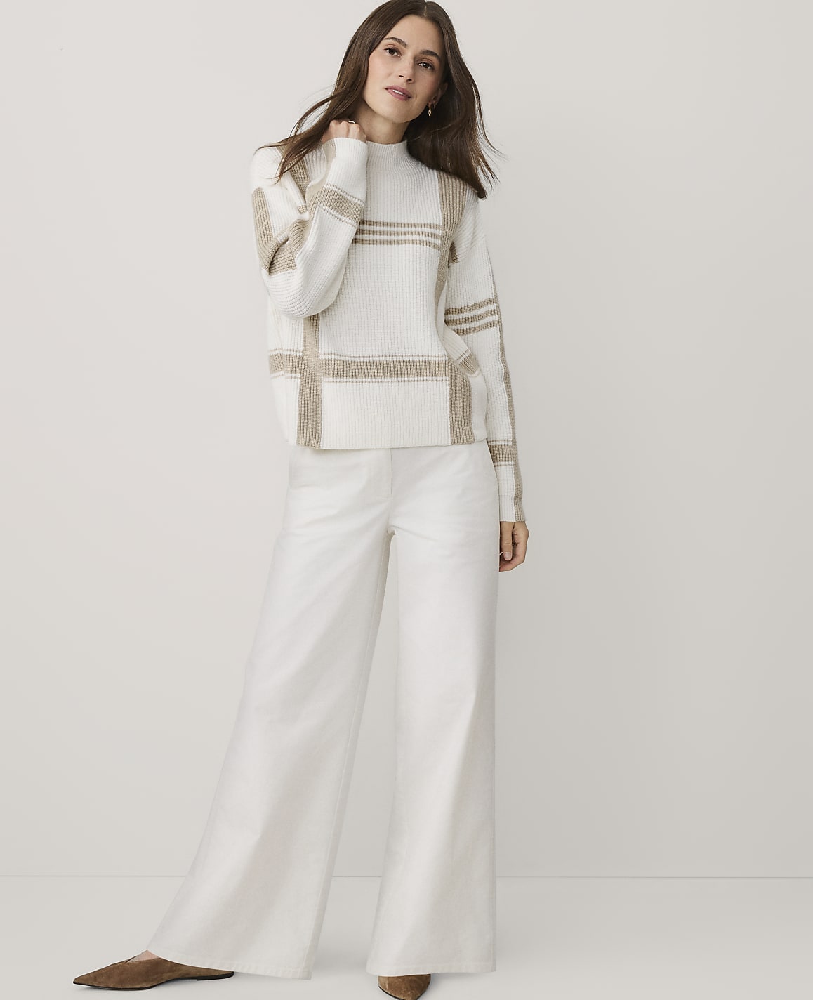 A woman wearing a beige and white patterned sweater and wide-leg white trousers stands against a plain white background.