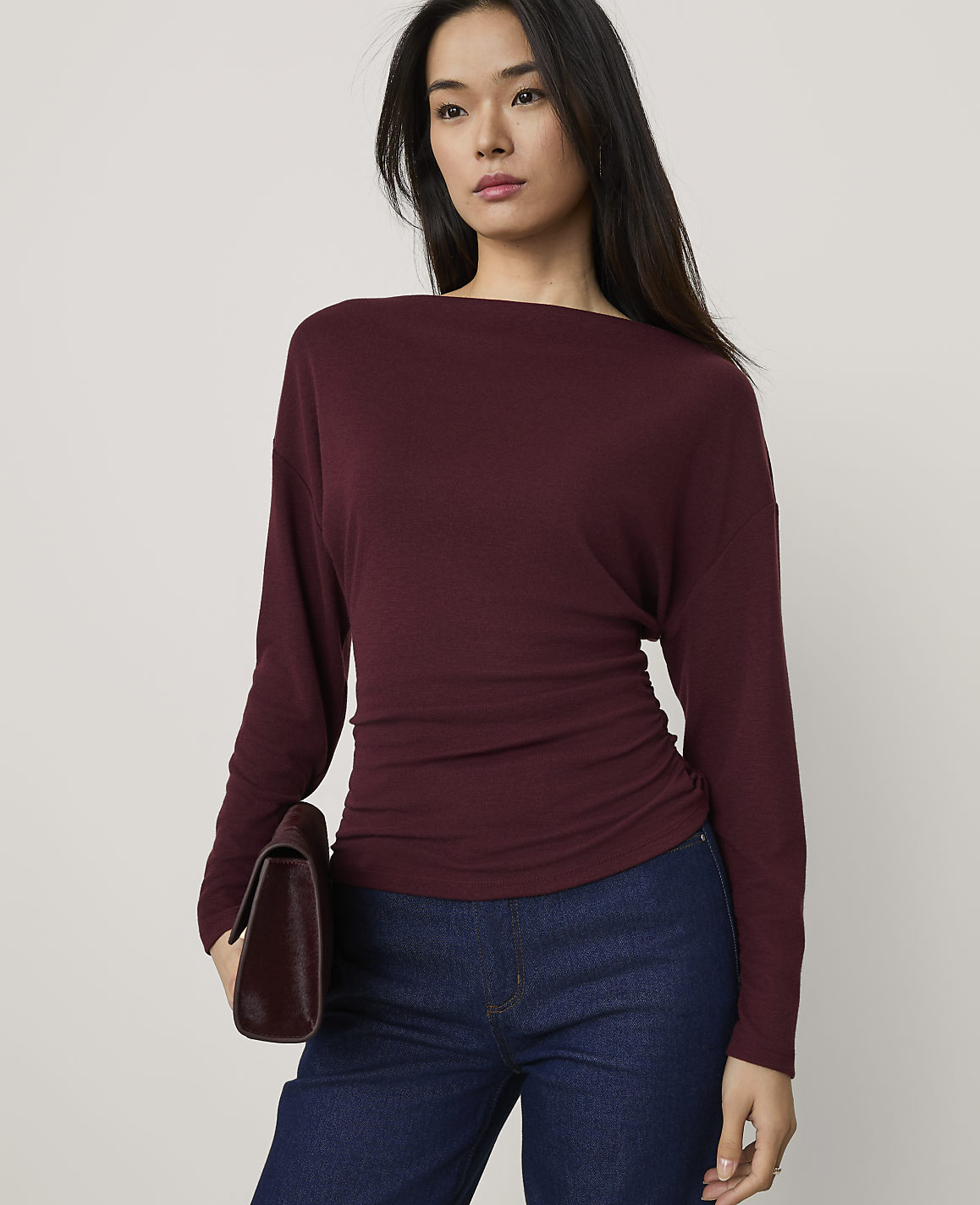 A woman wearing a burgundy long-sleeved top and blue jeans holds a dark clutch against a plain light gray background.