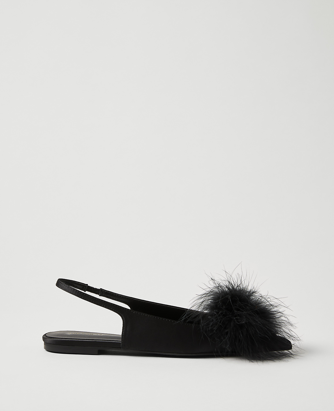 A black flat shoe with a fluffy pom-pom sits in the foreground against a plain, light gray background.