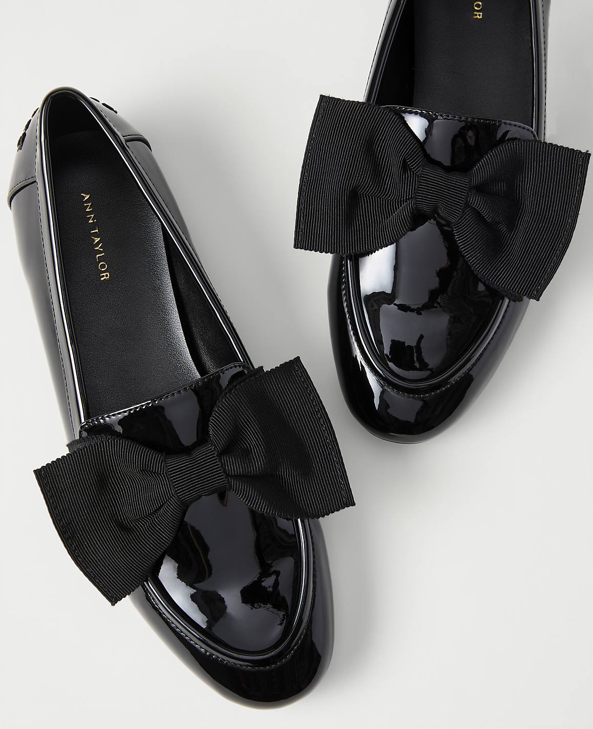 A pair of black patent leather loafers with large black bows are set against a plain white background.