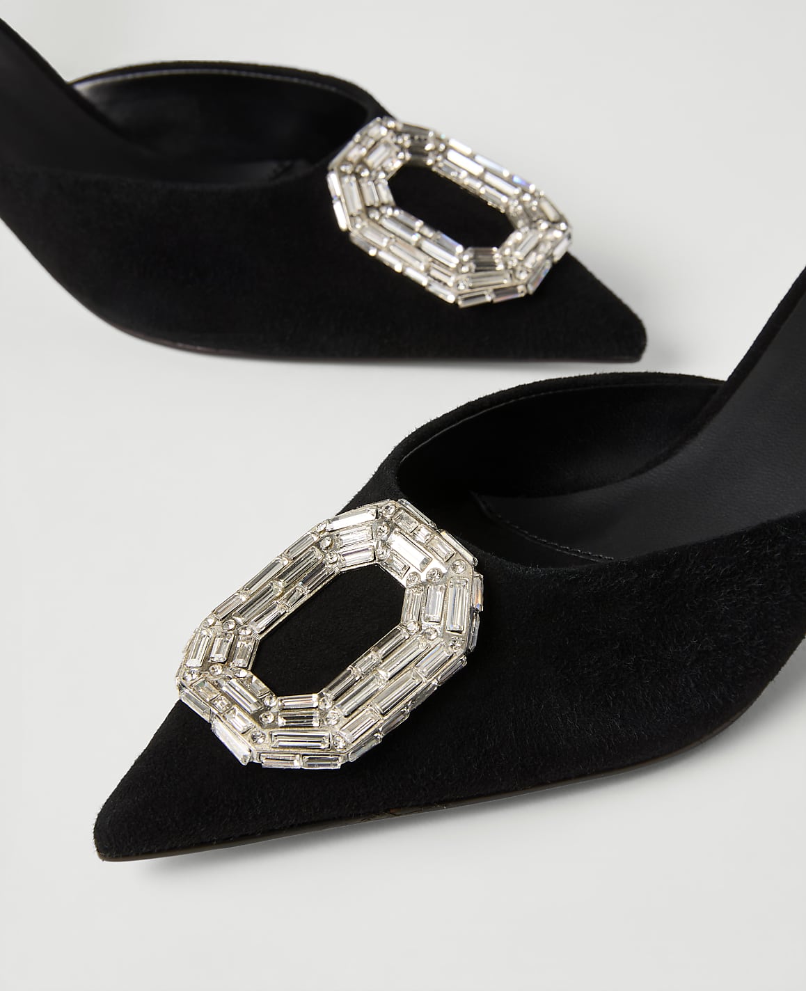Two black suede pointed-toe heels with large crystal buckles rest on a plain white background.