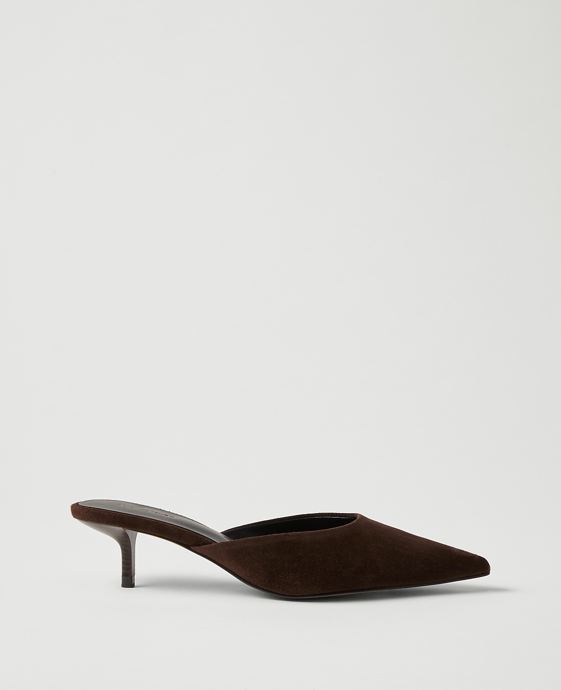 A brown suede pointed-toe mule heel sits against a plain, light gray background.