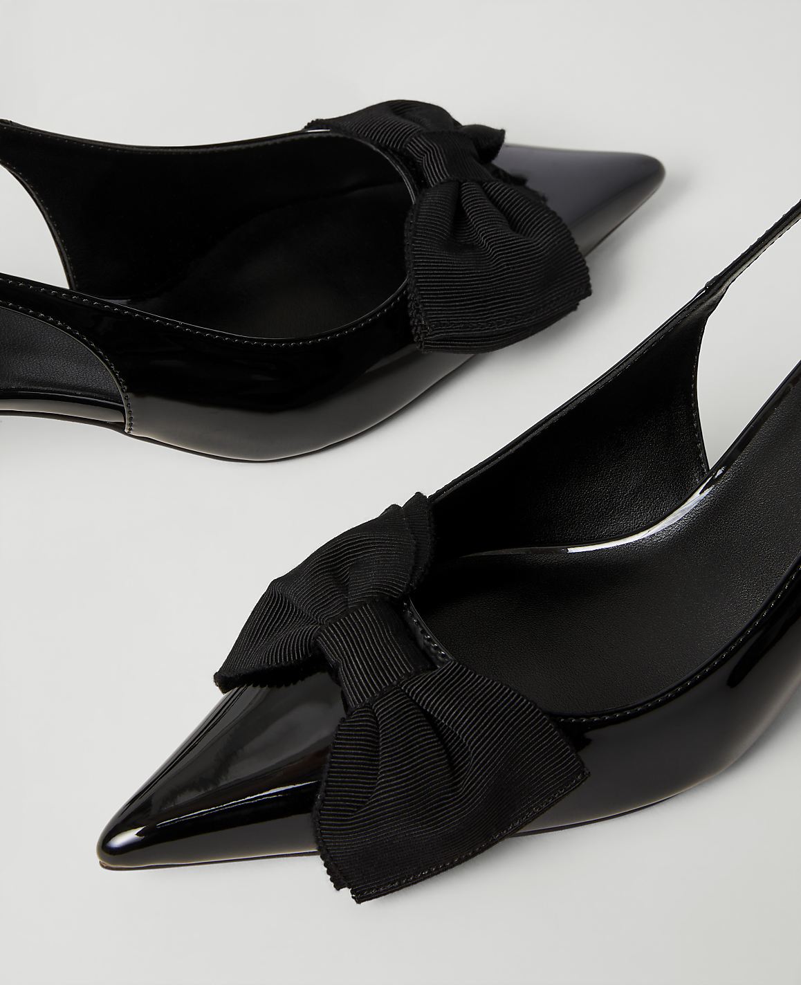 Two black patent leather pointed-toe heels with decorative bows sit against a plain white background.