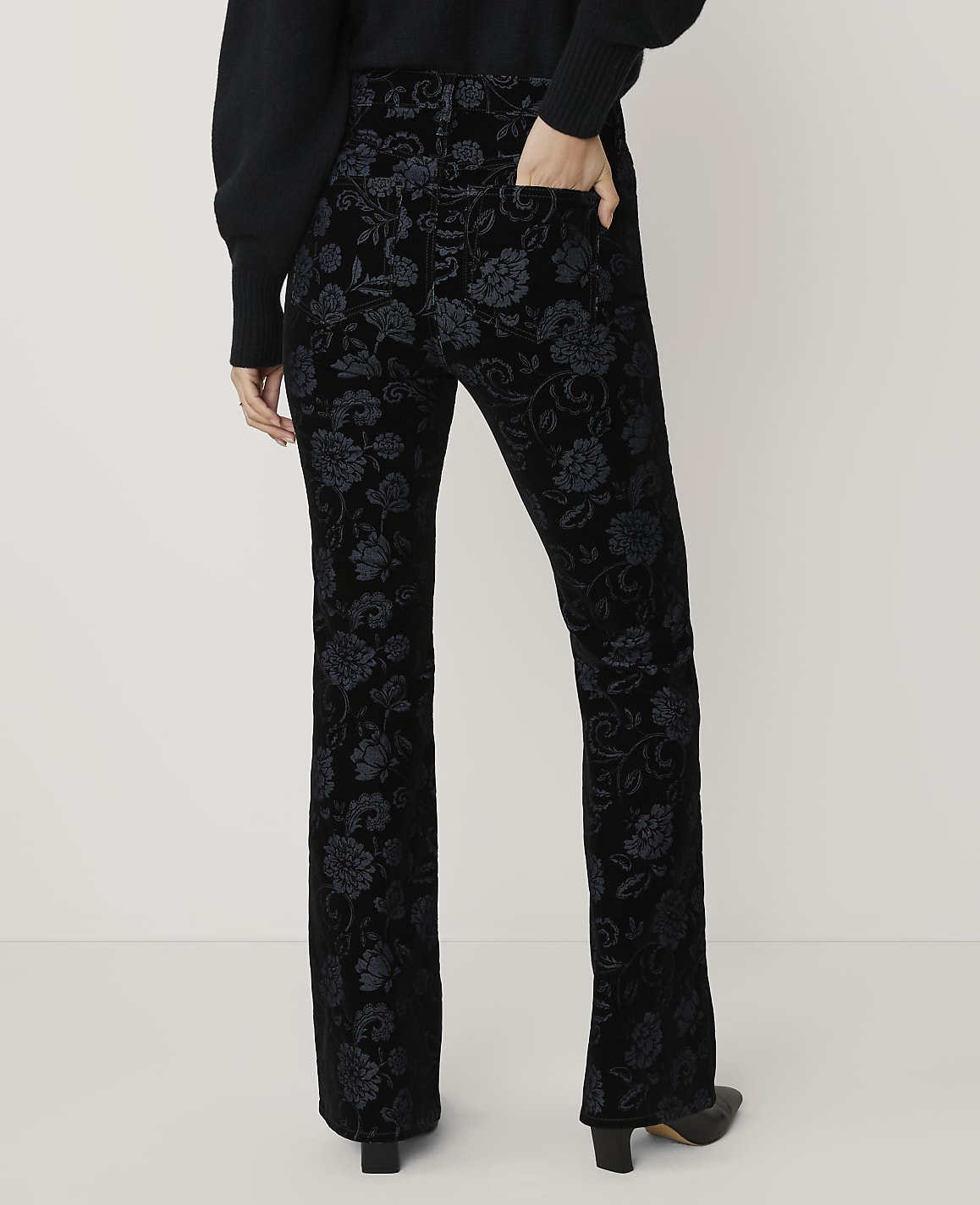 A person wears black floral patterned trousers and a dark sweater against a plain white background.