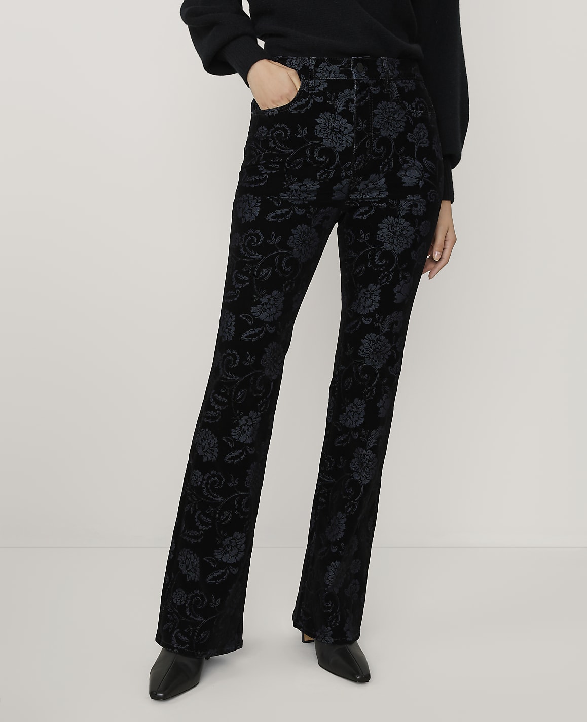 A person wearing a black sweater and floral-patterned black velvet trousers stands against a plain white background.