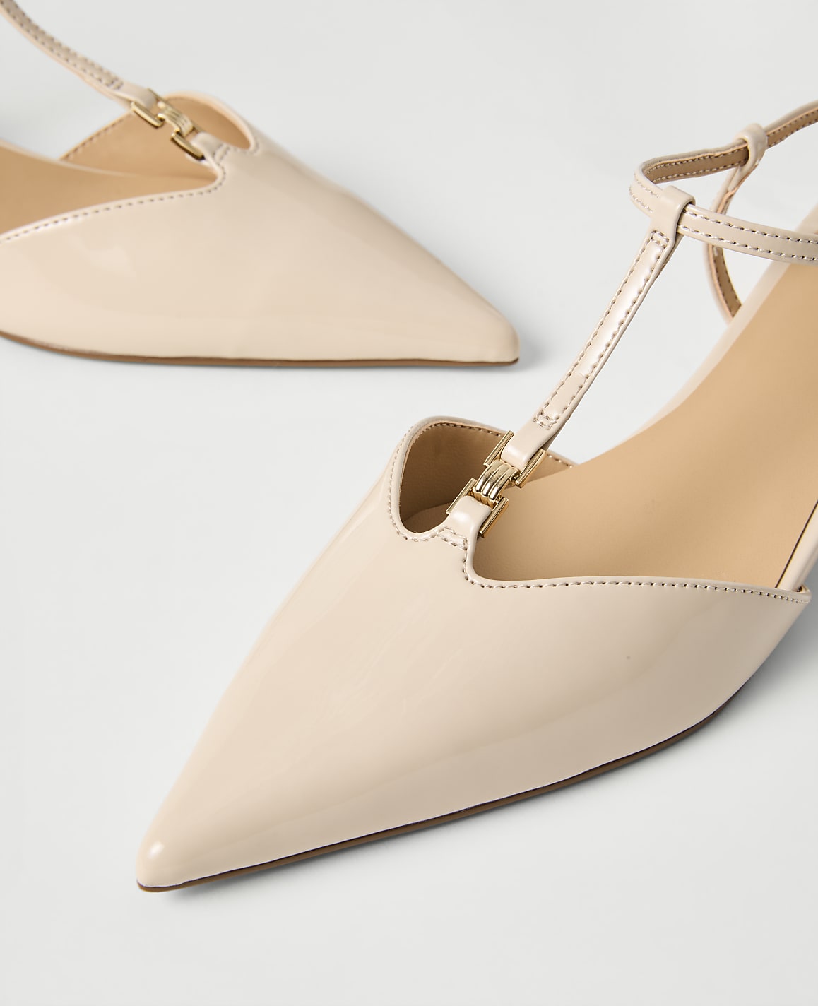 Two pointed-toe, beige patent leather heels with gold accents rest on a plain white surface.