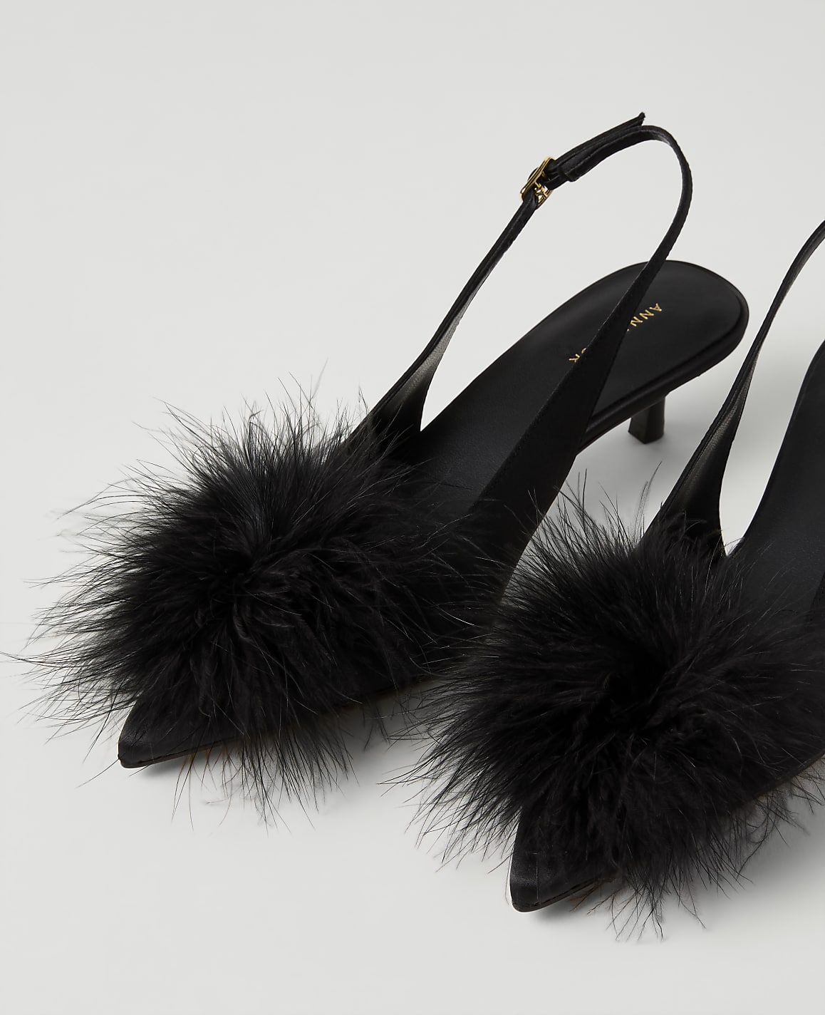 A pair of black pointed-toe heels with large fluffy pom-poms on the front are set against a plain white background.