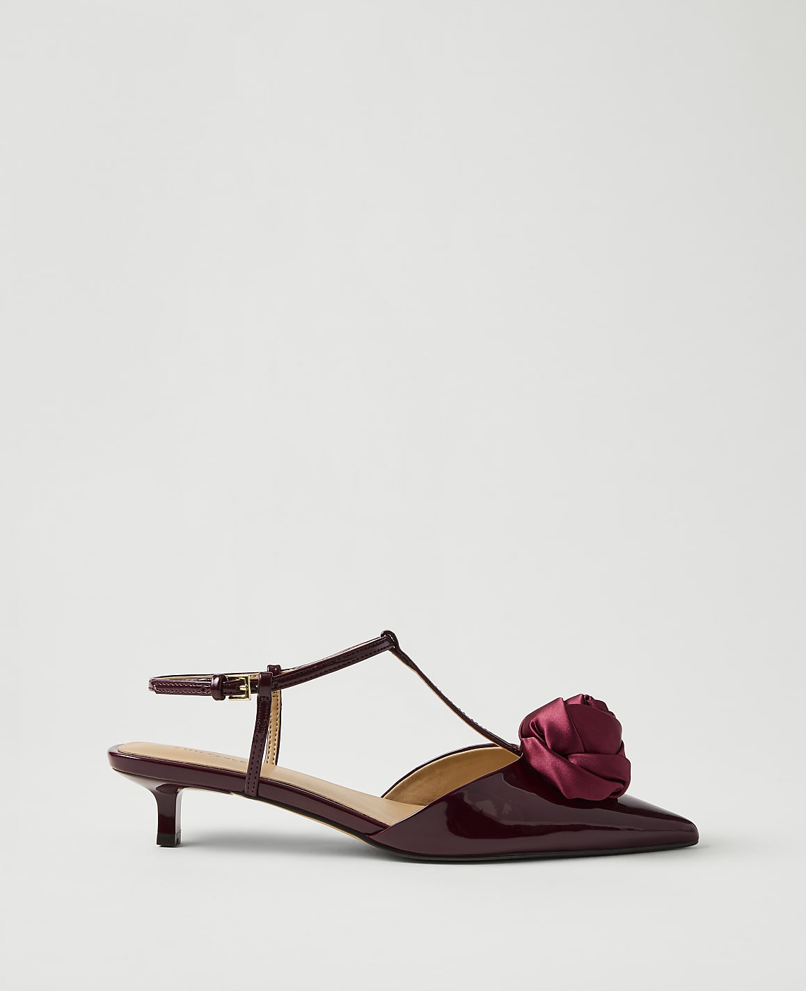 A burgundy patent leather kitten heel with a fabric rose detail sits against a plain, light grey background.