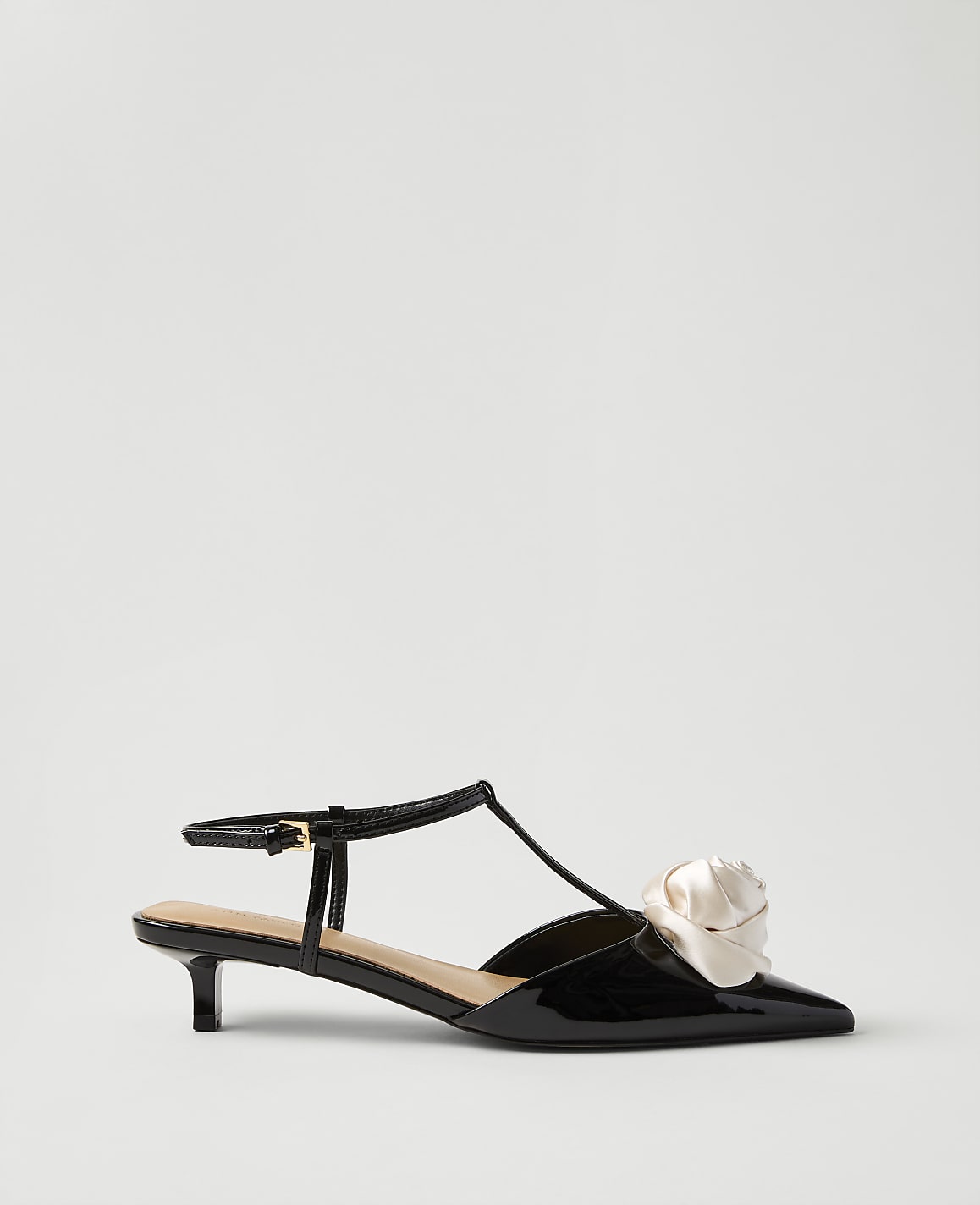 A black patent leather kitten heel with a cream-colored fabric flower on the toe sits against a plain, light gray background.