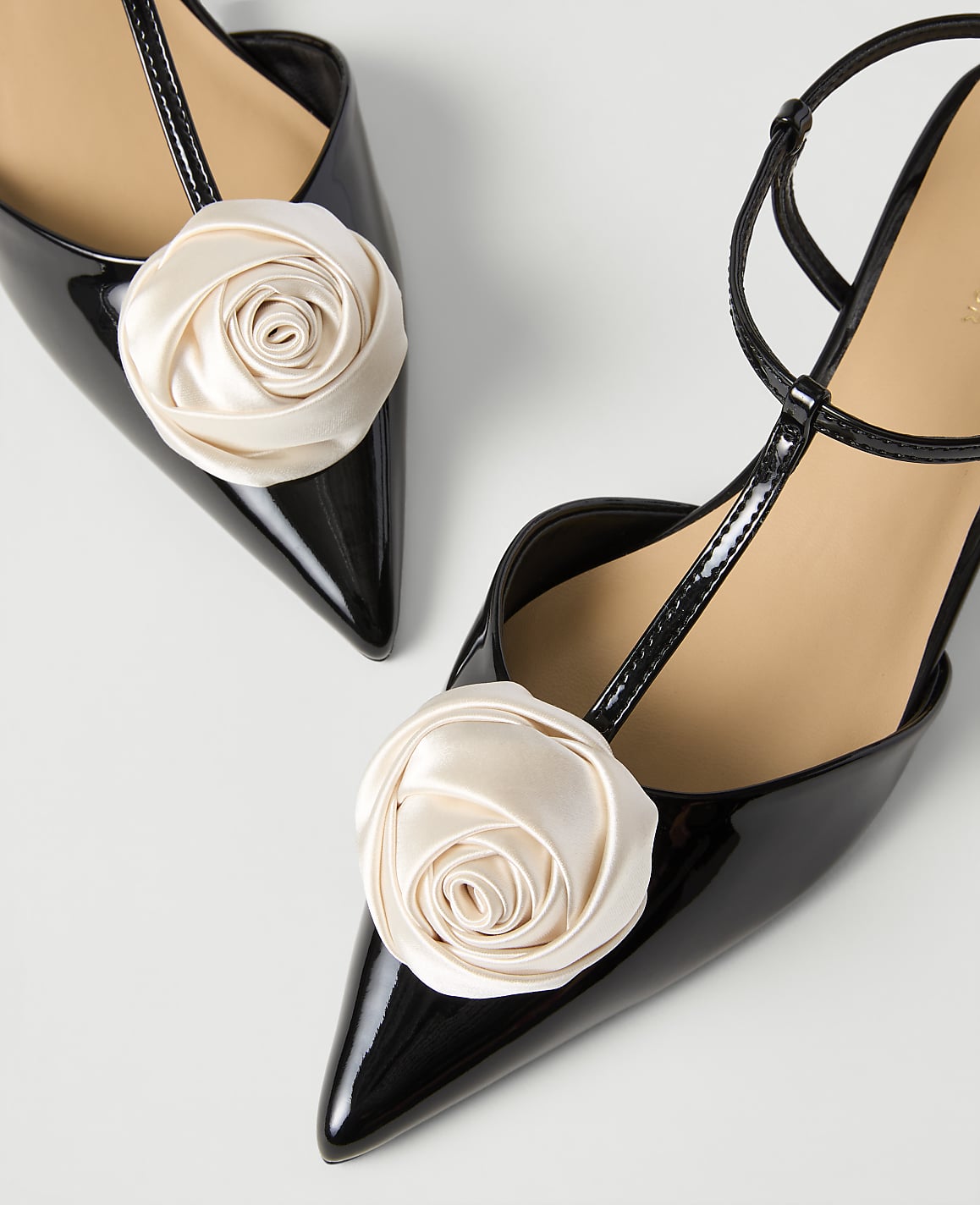 Two black patent leather pointed-toe heels with cream-colored fabric roses on the toes are set against a plain white background.
