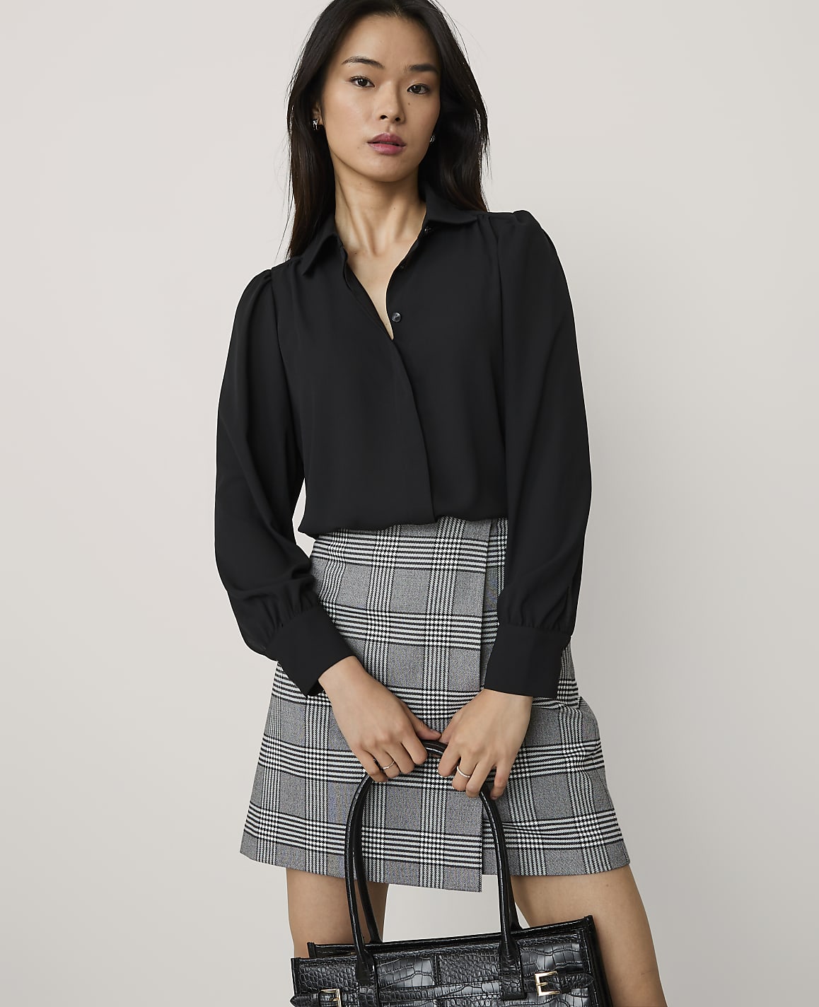 A woman wearing a black long-sleeved blouse and a grey plaid skirt holds a black handbag against a plain light grey background.