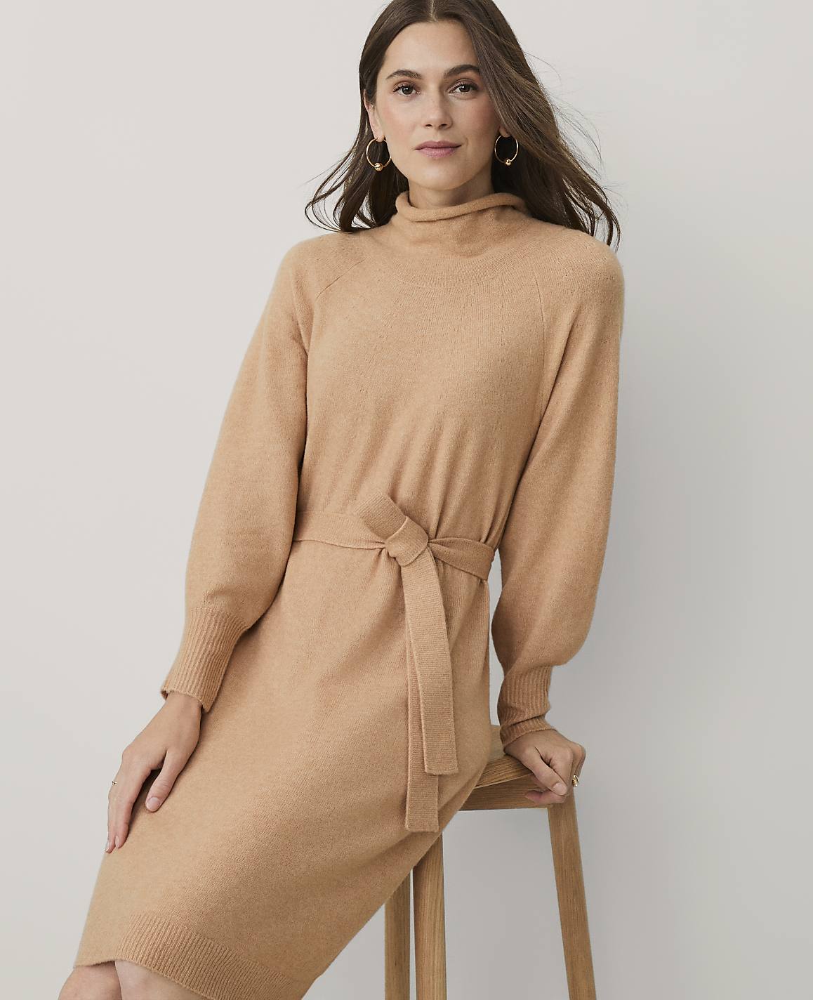 A woman wearing a camel-colored knit dress with a tie-waist sits on a wooden stool against a plain, light-colored background.