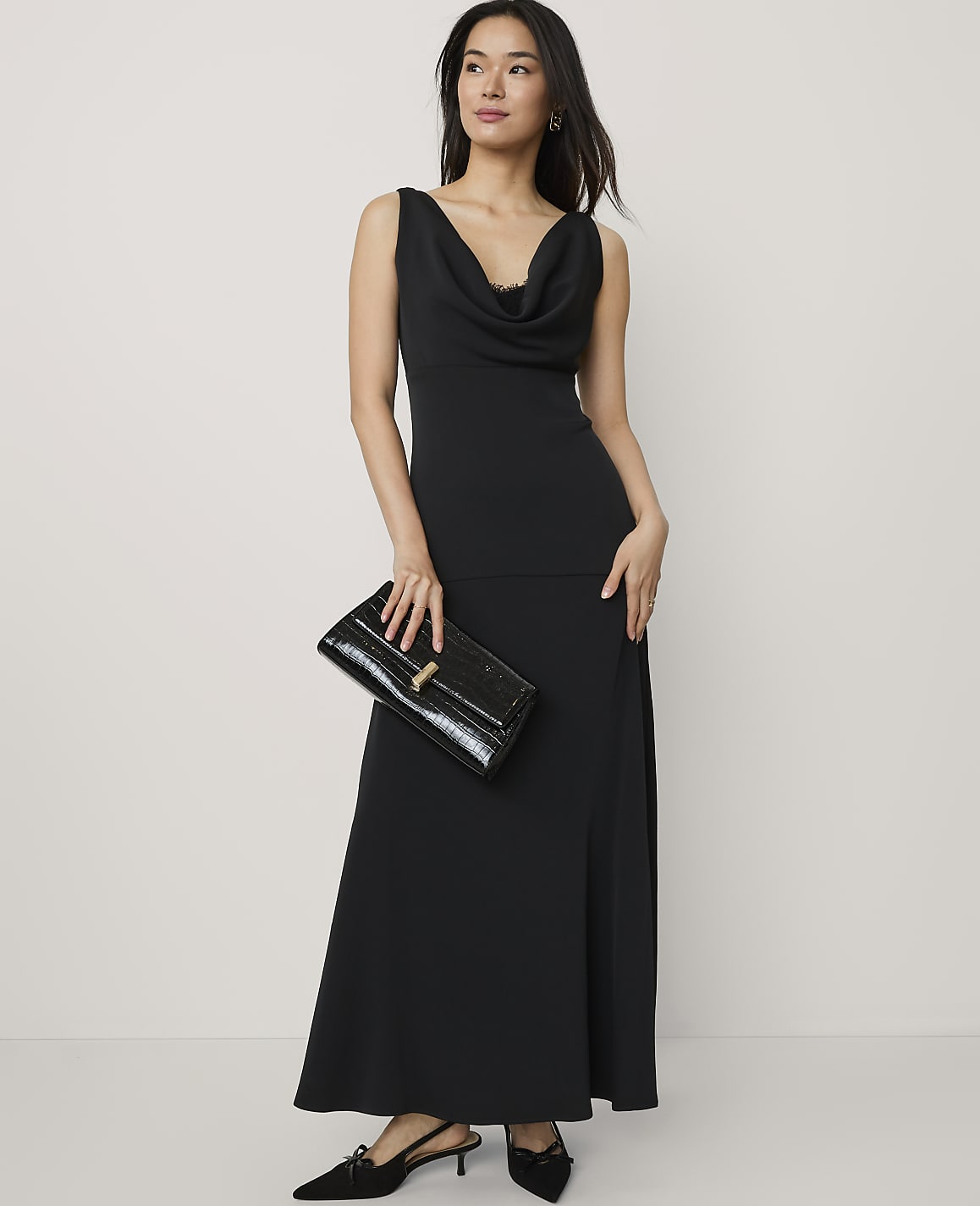 A woman wearing a long black cowl-neck dress and holding a black clutch stands against a plain, light-colored background.