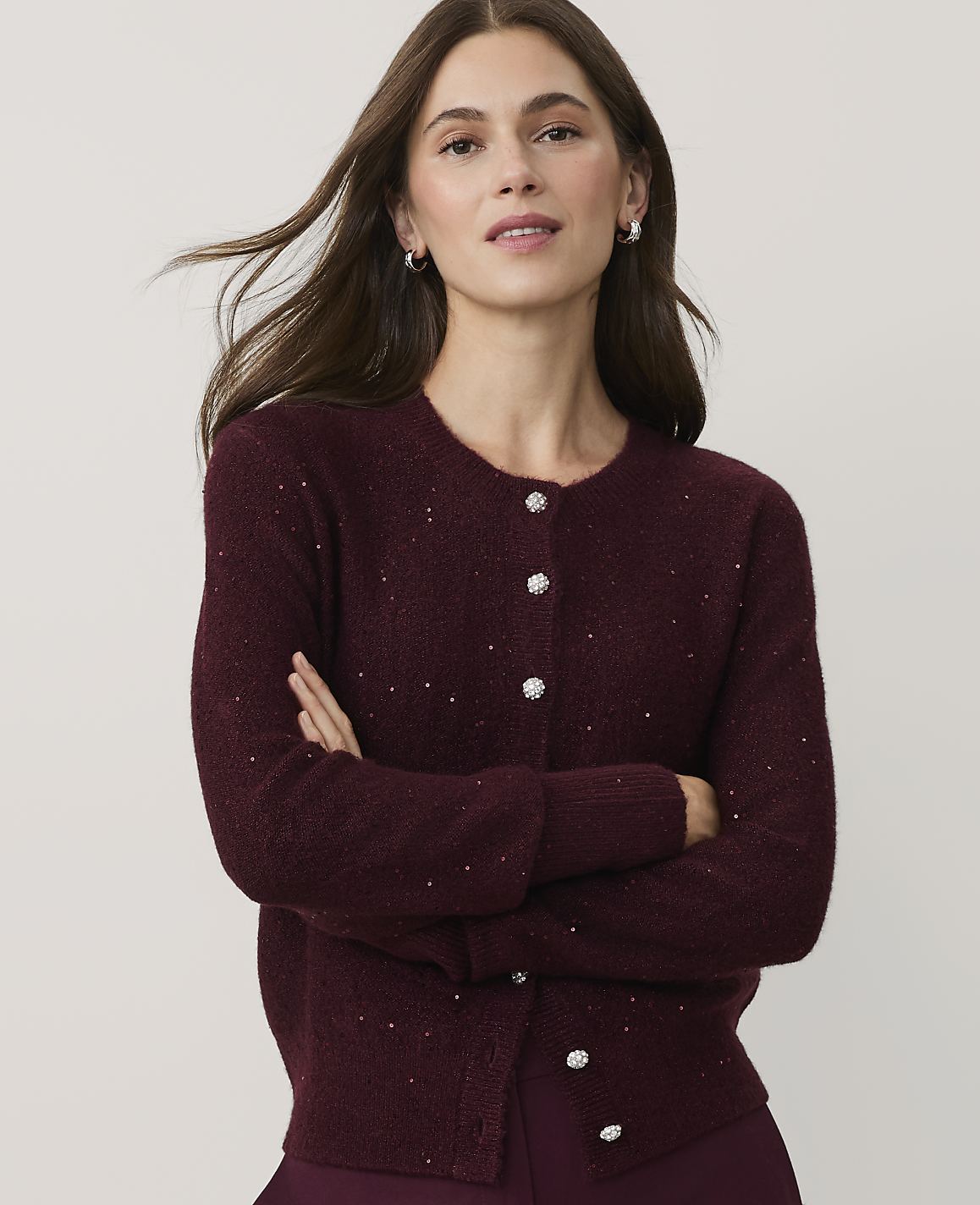 A woman with long brown hair wearing a sparkling burgundy cardigan stands with her arms crossed against a plain light gray background.