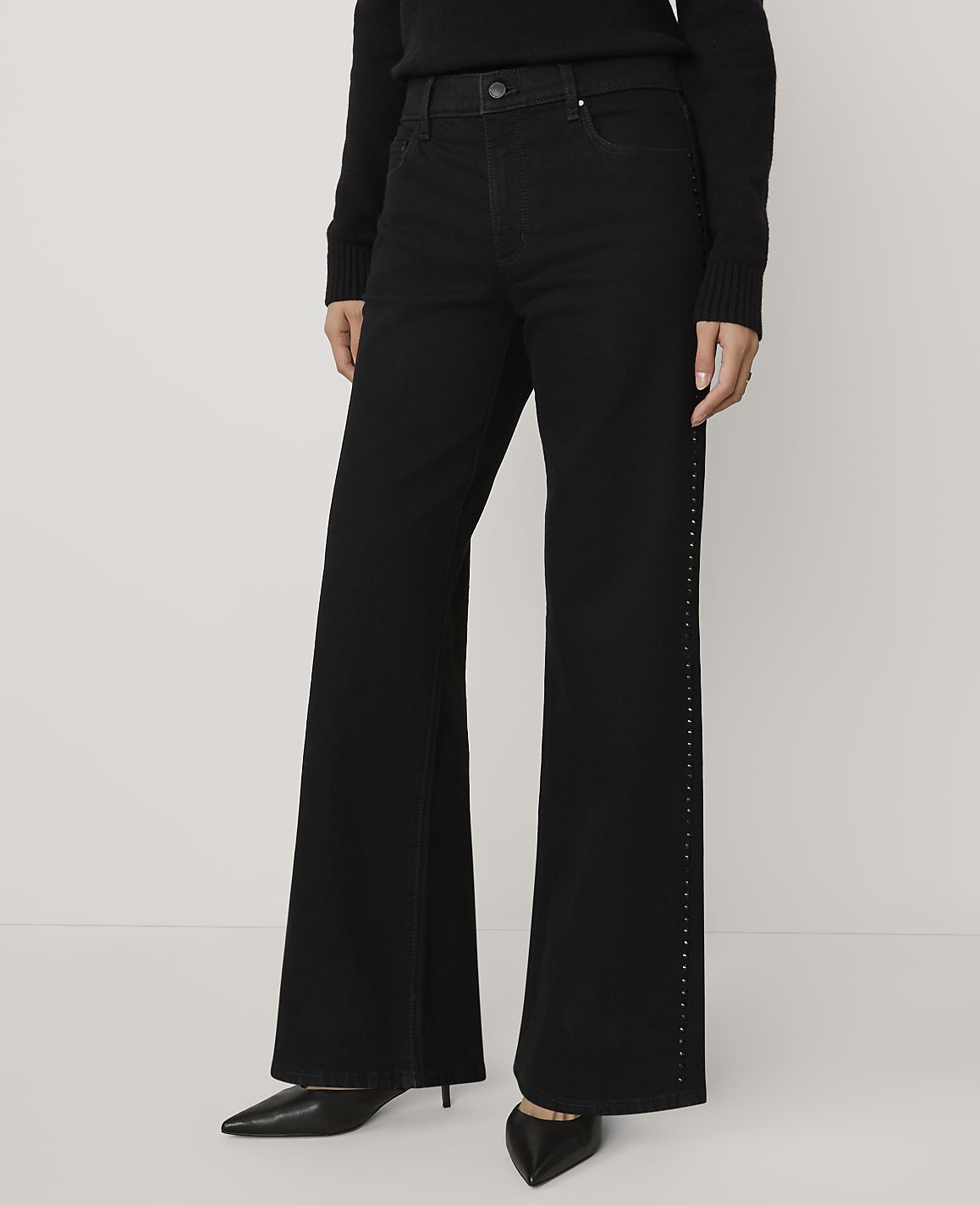 A person wearing a black sweater, wide-leg black trousers with side embellishments, and black pointed heels stands against a plain white background.