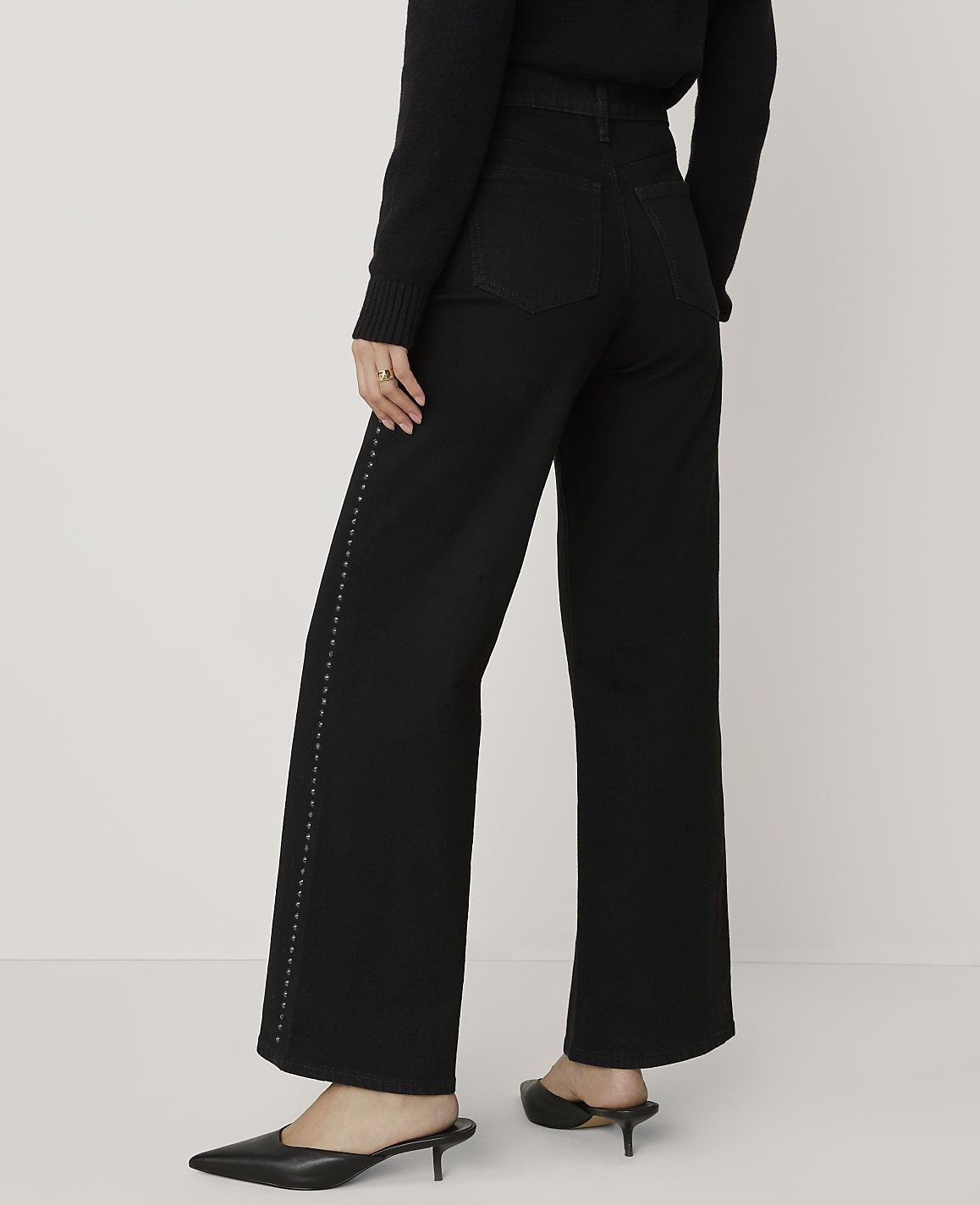 A person wearing a black sweater, wide-leg black trousers with side stud detailing, and black kitten heels stands against a plain white background.