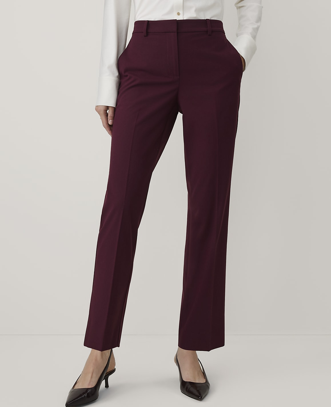 A person wearing a white button-down shirt, burgundy trousers, and black pointed heels stands against a plain white background.