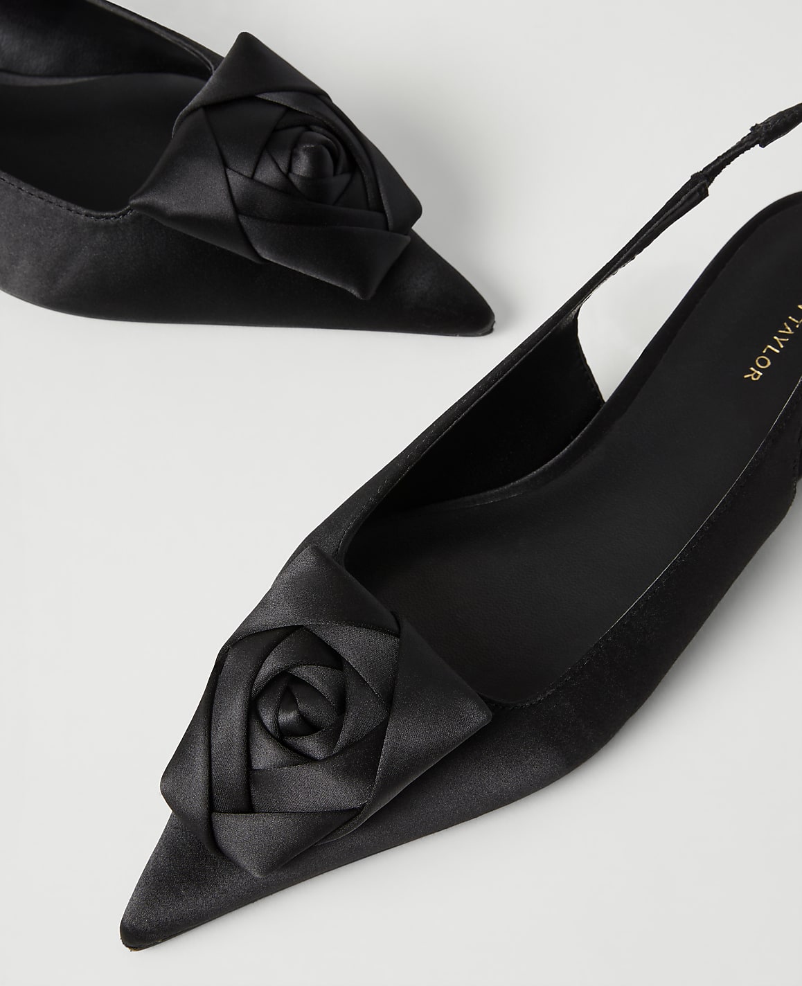 Two black satin pointed-toe heels with rose accents are positioned diagonally against a plain white background.