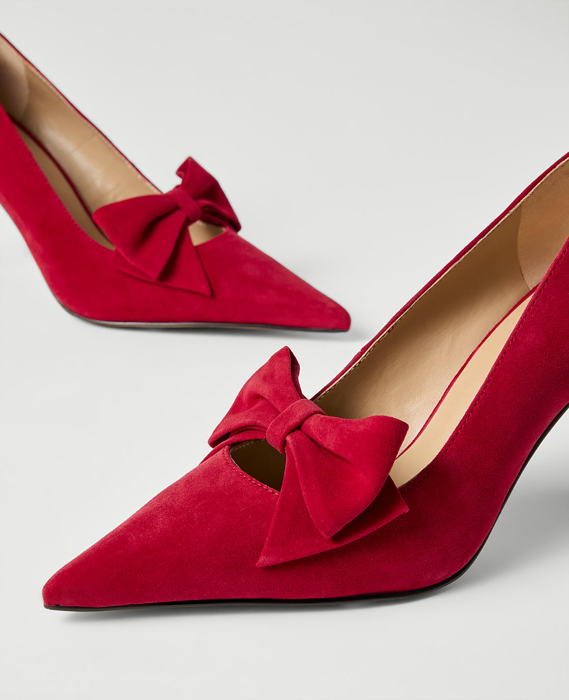 A pair of red suede pointed-toe pumps with decorative bows sit on a plain white background.