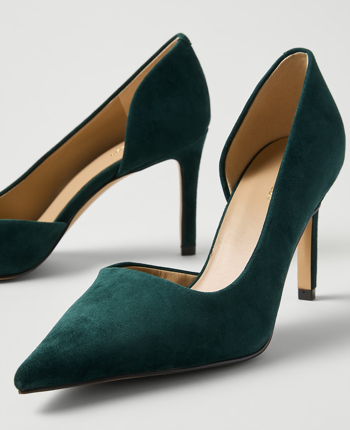A pair of dark green suede pointed-toe pumps sit against a plain light gray background.