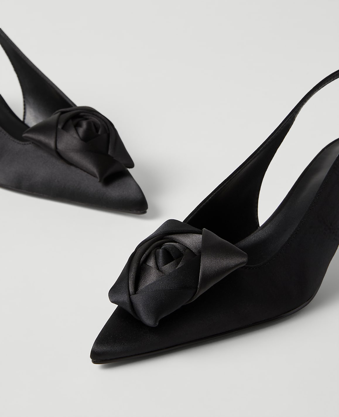 A pair of black satin slingback heels with rose-shaped ornaments on the toes are set against a plain light gray background.