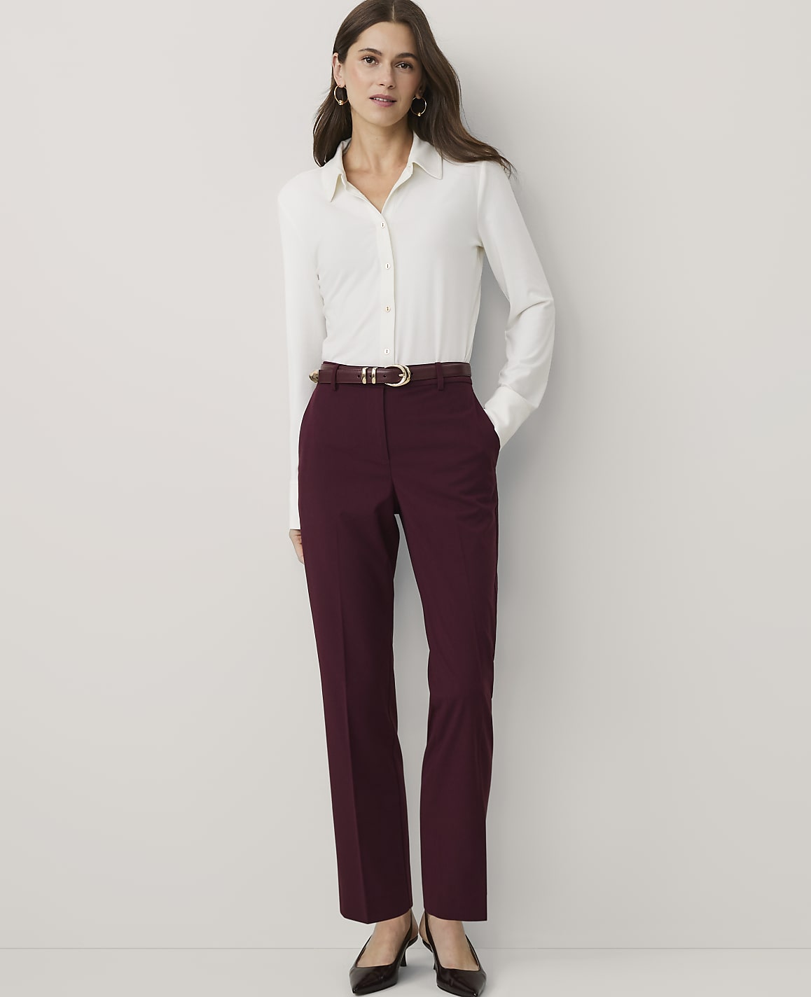 A woman wearing a white button-down shirt and burgundy trousers stands against a plain white background.