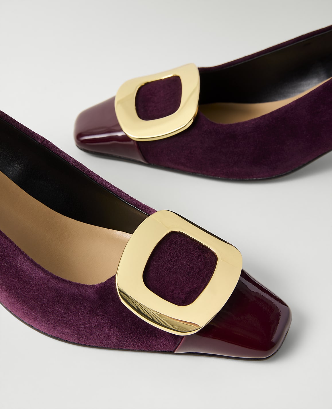 Two burgundy suede pumps with gold buckles and patent leather toes rest on a plain white background.